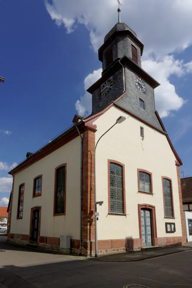 Die evangelische Kirche in Harreshausen, von der Straßenecke aus schräg von vorn: ein verputzter Saalbau mit rotem Sandstein an Ecken, Fenstergewänden und Portal, darüber ein achteckiger, ganz mit Schiefer gedeckter Turm mit zwei Zifferblättern und geschweifter Haube.