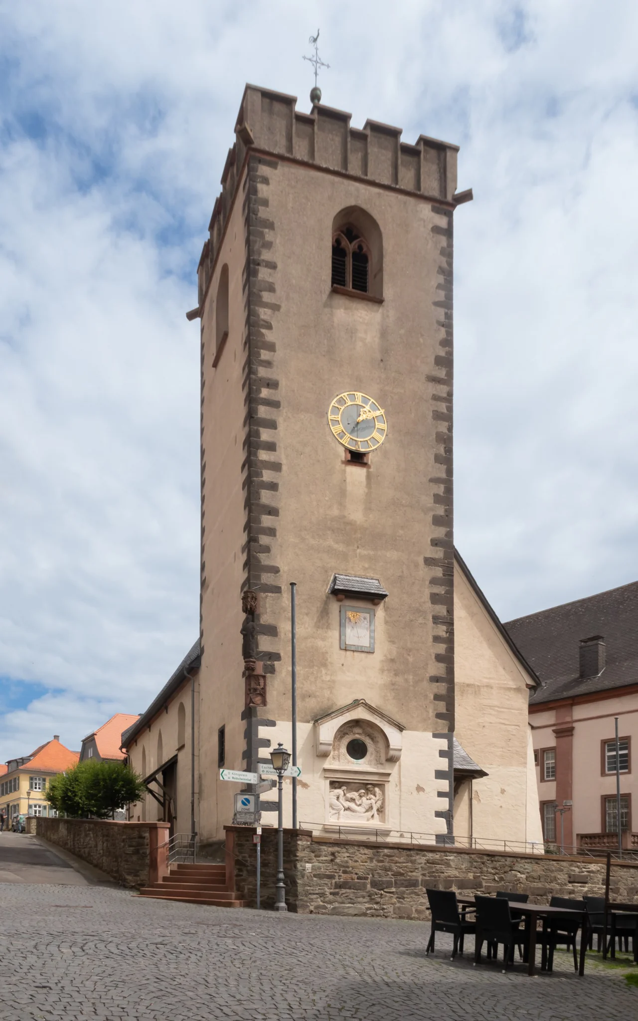 Der Turm der Stadtkirche St. Johann in Kronberg vom gepflasterten Platz aus, von Süden: ein hoher Bruchsteinturm mit Zinnenkranz und Wetterhahn, darin ein gotisches Maßwerkfenster, darunter die vergoldete Uhr, eine Sonnenuhr und über dem Portal ein Relief der Grablegung. Links schließt das niedrigere Langhaus mit Schieferdach an. Hochformat, 3000 × 4800 Pixel.
