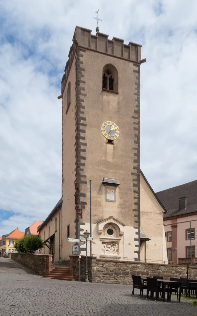 Der Turm der Stadtkirche St. Johann in Kronberg vom gepflasterten Platz aus, von Süden: ein hoher Bruchsteinturm mit Zinnenkranz und Wetterhahn, darin ein gotisches Maßwerkfenster, darunter die vergoldete Uhr, eine Sonnenuhr und über dem Portal ein Relief der Grablegung. Links schließt das niedrigere Langhaus mit Schieferdach an. Hochformat, 3000 × 4800 Pixel.