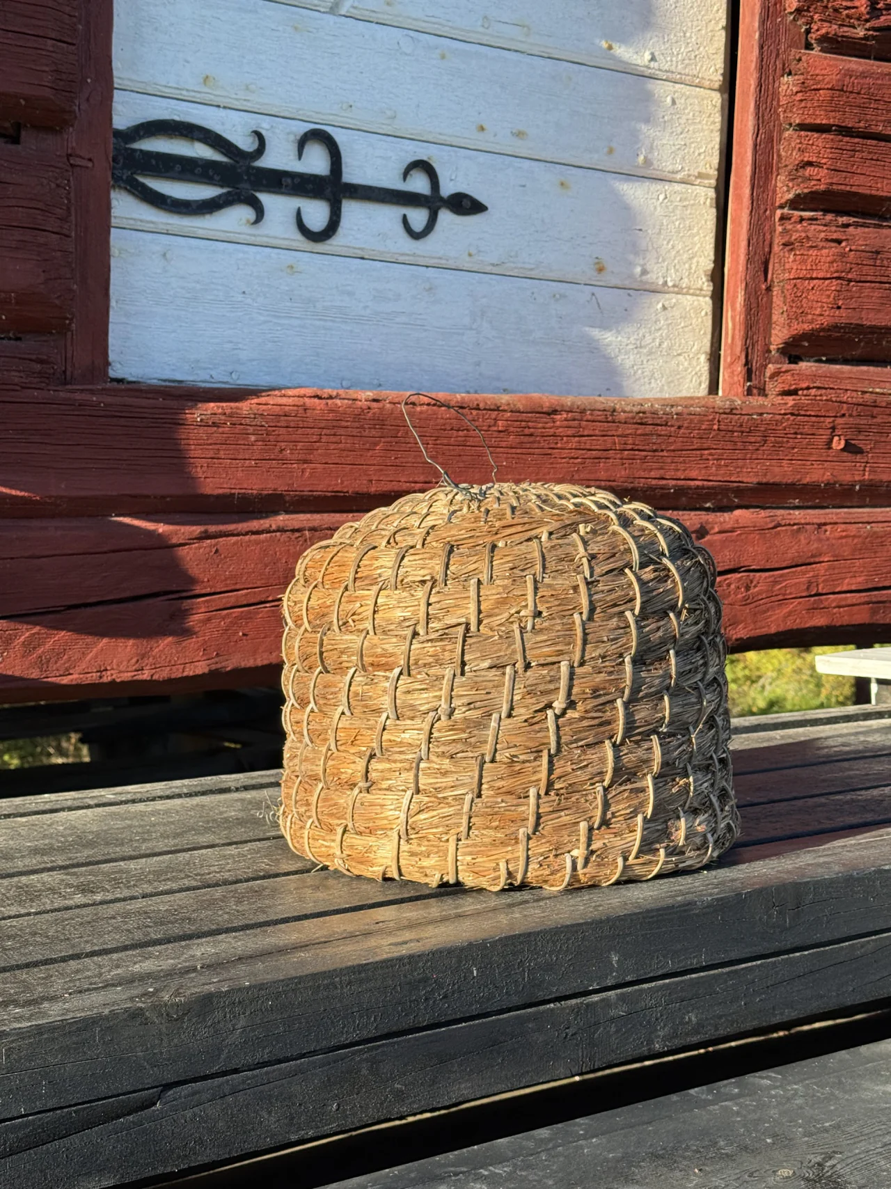 Stülpkorb aus gebundenen Strohwülsten auf einer verwitterten Holzbank vor einer roten Holzwand.